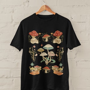 Könnte beinhalten: Schwarzes T-Shirt mit einem bunten Pilz-Design. Die Pilze sind in verschiedenen Formen und Größen, einige haben rote Hüte. Das Design ist von grünen Ranken und Sternen umgeben.