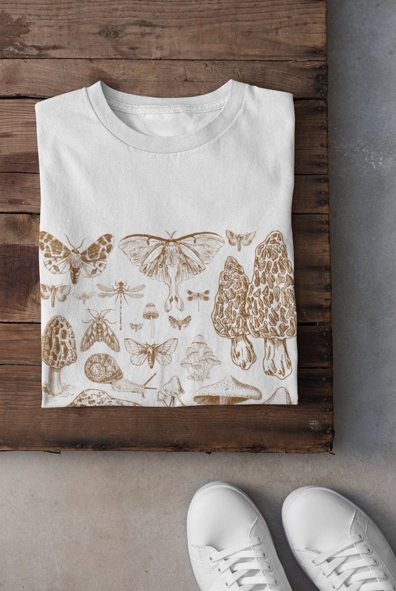 K&ouml;nnte beinhalten: Ein wei&szlig;es T-Shirt mit einem braunen Druck verschiedener Insekten und Pilze. Der Druck umfasst Schmetterlinge, Motten, Libellen, Schnecken und Pilze.