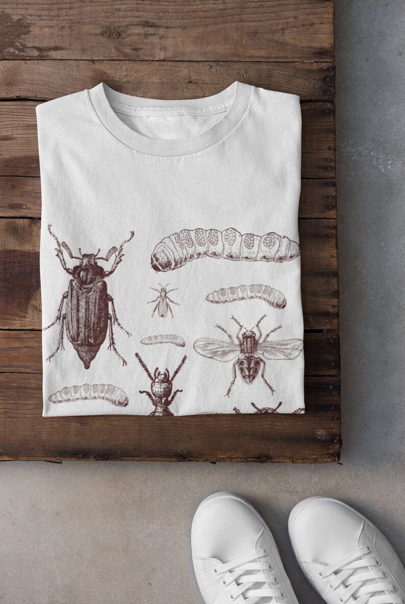 May include: White t-shirt with a brown illustration of insects, including beetles, caterpillars, and a fly. The shirt is folded on a wooden surface, with white trainers visible. The design has a vintage, scientific aesthetic, with the shirt folded.
