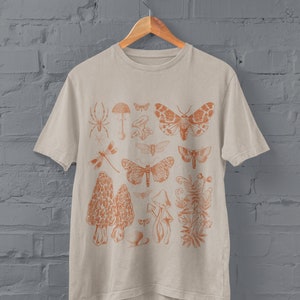 May include: A beige t-shirt featuring an orange print of various insects and mushrooms. The design includes a spider, dragonfly, moths, mushrooms, and a plant.