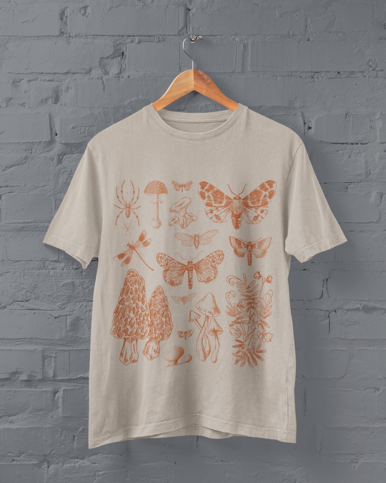 May include: A beige t-shirt featuring an orange print of various insects and mushrooms. The design includes a spider, dragonfly, moths, mushrooms, and a plant.