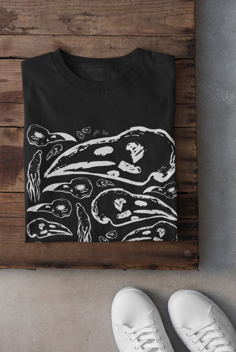 Op de afbeelding: Zwart T-shirt met een witte grafische afbeelding van vogel schedels en abstracte vormen. Het shirt is opgevouwen op een houten oppervlak, met witte sneakers zichtbaar aan de onderkant. Een casual, gothic-ge&iuml;nspireerd kledingstuk.