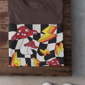 May include: A brown t-shirt with a checkered pattern featuring red mushrooms with eyes and yellow lightning bolts.