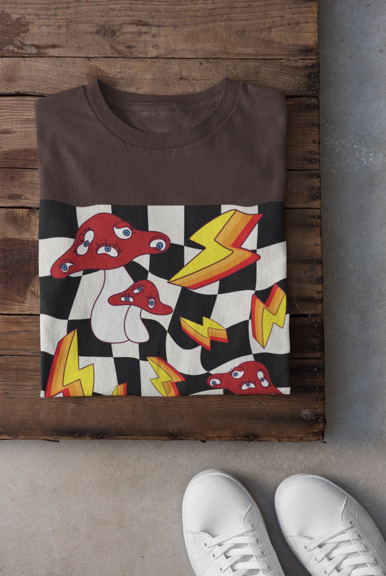 May include: A brown t-shirt with a checkered pattern featuring red mushrooms with eyes and yellow lightning bolts.