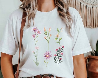 Può includere: Maglietta bianca con stampa floreale con fiori rosa, bianchi e viola. I fiori sono disposti in modo simmetrico sul davanti della maglietta.
