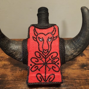 Könnte beinhalten: Ein roter Filz-Flaschenüberzug mit einem schwarzen Bullendesign und schwarzen Strasssteinen. Der Flaschenüberzug ist mit schwarzem Garn umwickelt und an einem Paar braunen Bullenhörnern befestigt.
