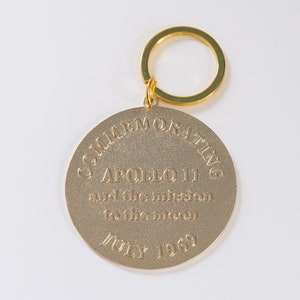May include: Gold-colored metal keychain commemorating Apollo 11 and the mission to the moon. The keychain features the text "COMMEMORATING APOLLO 11 and the mission to the moon JULY 1969".