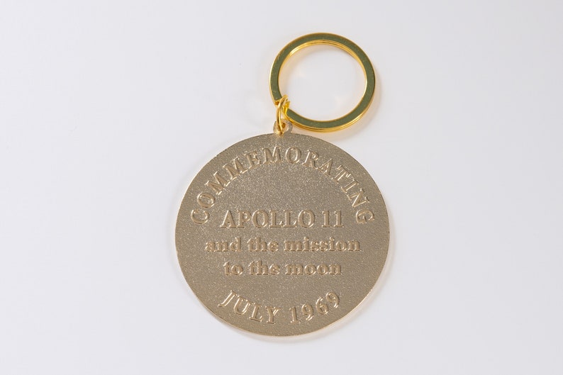 May include: Gold-colored metal keychain commemorating Apollo 11 and the mission to the moon. The keychain features the text "COMMEMORATING APOLLO 11 and the mission to the moon JULY 1969".