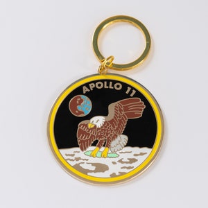 May include: A gold-colored keychain with a round black and white enamel charm featuring the Apollo 11 mission logo. The logo depicts an eagle landing on the moon with the Earth in the background and the text "APOLLO 11".