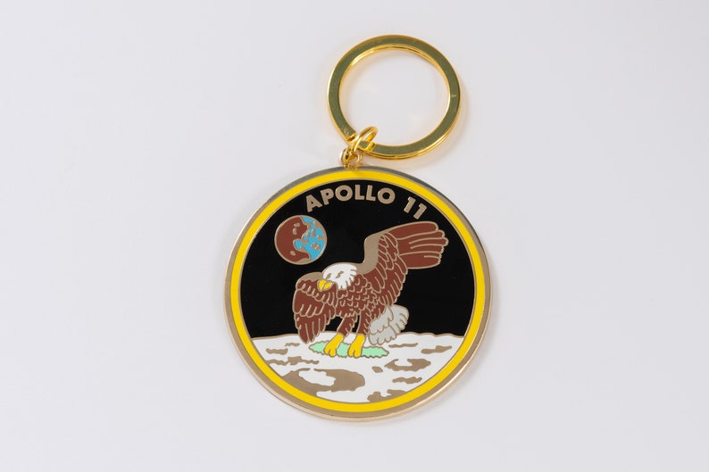 May include: A gold-colored keychain with a round black and white enamel charm featuring the Apollo 11 mission logo. The logo depicts an eagle landing on the moon with the Earth in the background and the text "APOLLO 11".