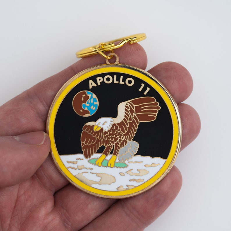 May include: A gold-colored keychain with a round, black enamel charm featuring a bald eagle landing on the moon. The charm has a yellow border and the text "APOLLO 11" in white lettering.