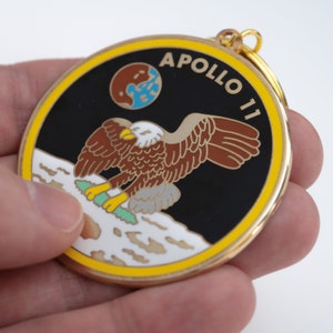 May include: A gold-colored metal keychain with a black and white image of an eagle landing on the moon. The keychain has the text "APOLLO 11" in a yellow circle around the image.