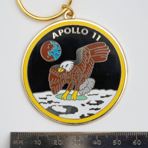 May include: A gold-colored keychain with a black and white enamel design. The design features an eagle landing on the moon with the words "Apollo 11" in a yellow banner.