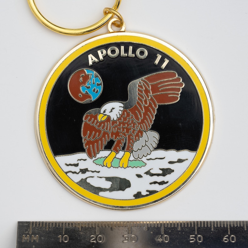 May include: A gold-colored keychain with a black and white enamel design. The design features an eagle landing on the moon with the words "Apollo 11" in a yellow banner.
