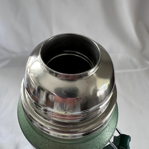 May include: A green vacuum-insulated thermos with a textured finish and a polished stainless steel rim at the top opening. The container features a threaded neck for a cap and a dark green plastic handle attached to the side of the cylindrical body.