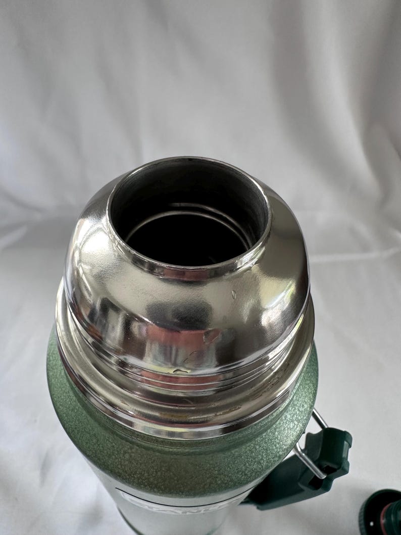 May include: A green vacuum-insulated thermos with a textured finish and a polished stainless steel rim at the top opening. The container features a threaded neck for a cap and a dark green plastic handle attached to the side of the cylindrical body.