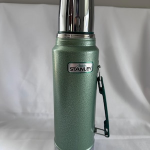 May include: A green, textured, cylindrical vacuum-insulated thermos with a shiny silver cap and base. The body features a rectangular emblem with the text Aladdin STANLEY. A dark green plastic handle is attached to the side of the bottle.