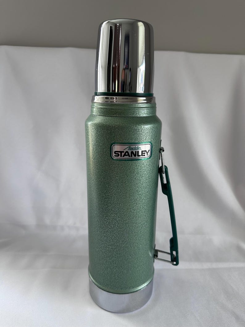 May include: A green, textured, cylindrical vacuum-insulated thermos with a shiny silver cap and base. The body features a rectangular emblem with the text Aladdin STANLEY. A dark green plastic handle is attached to the side of the bottle.