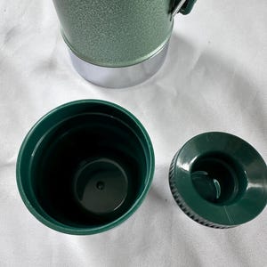 May include: A green vacuum insulated bottle with a textured finish and a silver base. The bottle features a dark green plastic cup lid and a separate dark green screw-on stopper. A green handle is attached to the side of the bottle body.