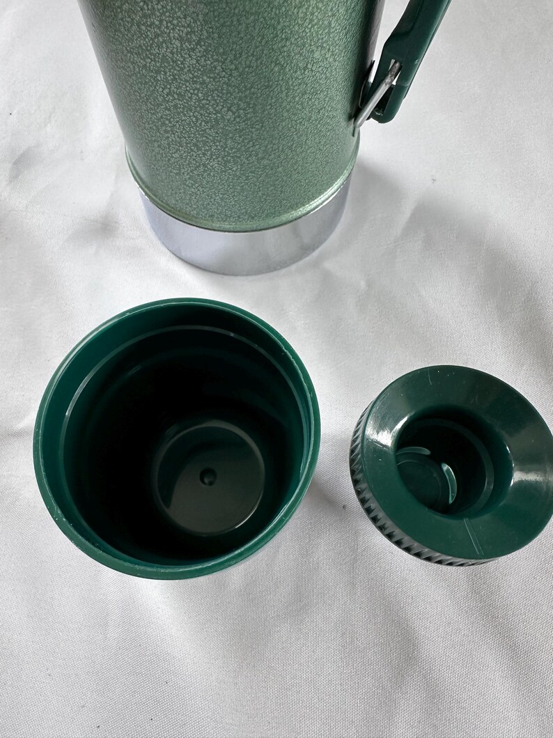 May include: A green vacuum insulated bottle with a textured finish and a silver base. The bottle features a dark green plastic cup lid and a separate dark green screw-on stopper. A green handle is attached to the side of the bottle body.