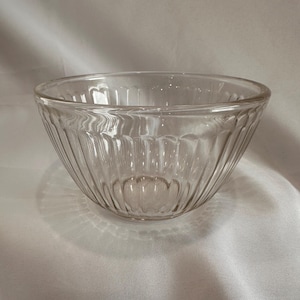 May include: A clear glass bowl with vertical ribbed detailing. The bowl has a rounded shape and a scalloped base. The glass is transparent, allowing light to pass through. The bowl is likely used for serving or displaying food items.