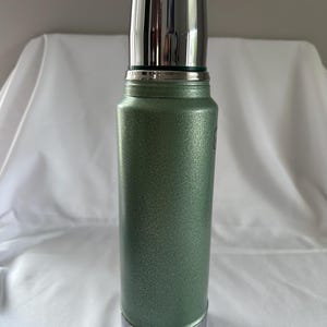 May include: A cylindrical vacuum-insulated thermos featuring a textured, sage green metallic finish. The container includes a polished, reflective silver-toned cap that functions as a cup, along with a matching silver-toned base at the bottom.