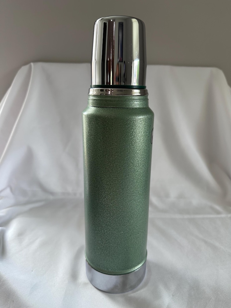 May include: A cylindrical vacuum-insulated thermos featuring a textured, sage green metallic finish. The container includes a polished, reflective silver-toned cap that functions as a cup, along with a matching silver-toned base at the bottom.