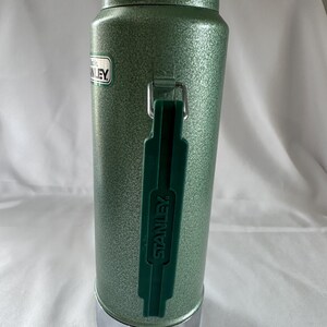 May include: A sage green Stanley vacuum insulated thermos with a textured hammer-tone finish. The cylindrical bottle features a dark green plastic handle with the word STANLEY embossed on it, a silver metallic base, and a silver metallic cap.