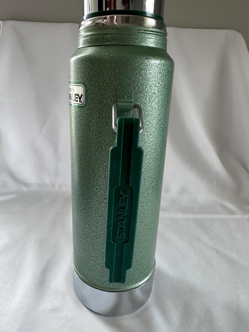 May include: A sage green Stanley vacuum insulated thermos with a textured hammer-tone finish. The cylindrical bottle features a dark green plastic handle with the word STANLEY embossed on it, a silver metallic base, and a silver metallic cap.