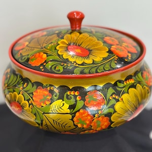 May include: A round wooden bowl with a matching lid featuring a red knob. The exterior is painted with a black background, vibrant yellow sunflowers, orange berries, and green leaves. The rim of the lid and the bowl are painted in a solid red color.