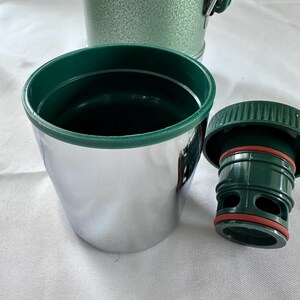 May include: A stainless steel cup with a dark green rim, accompanied by a matching dark green plastic stopper featuring a red silicone gasket. In the background, a portion of a textured, light green thermos container is visible.