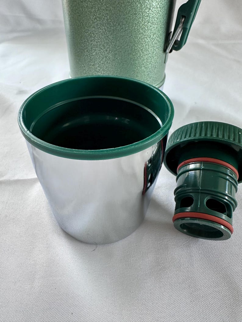 May include: A stainless steel cup with a dark green rim, accompanied by a matching dark green plastic stopper featuring a red silicone gasket. In the background, a portion of a textured, light green thermos container is visible.