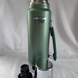 May include: A green, textured Stanley Aladdin vacuum bottle with a silver metallic cap and a matching silver cup. The bottle features a dark green folding handle on the side and a black and green stopper with a red rubber seal ring at the base.