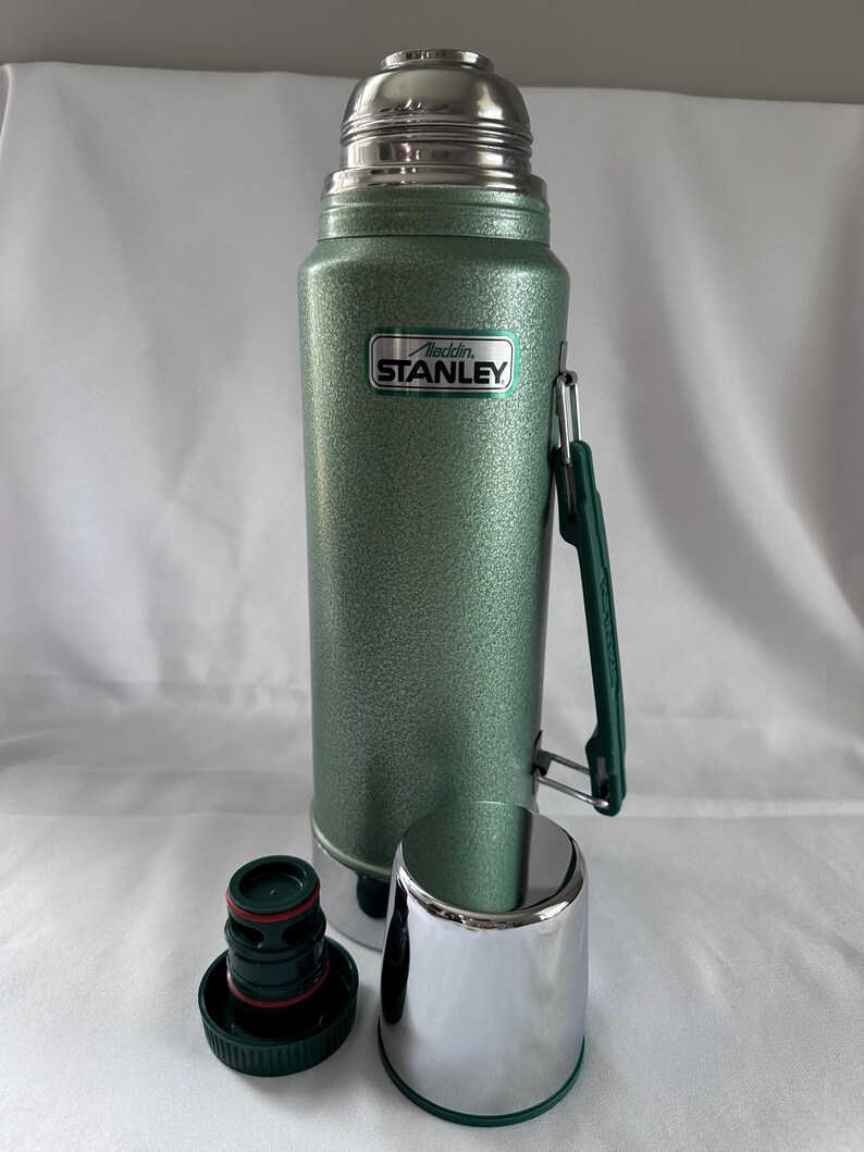 May include: A green, textured Stanley Aladdin vacuum bottle with a silver metallic cap and a matching silver cup. The bottle features a dark green folding handle on the side and a black and green stopper with a red rubber seal ring at the base.