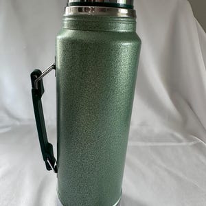 May include: A tall, cylindrical vacuum-insulated thermos with a textured, sage green hammered finish. It features a polished silver-tone metal cap and base, and a dark green plastic side handle attached with silver-tone metal hardware.