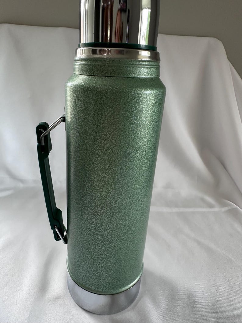 May include: A tall, cylindrical vacuum-insulated thermos with a textured, sage green hammered finish. It features a polished silver-tone metal cap and base, and a dark green plastic side handle attached with silver-tone metal hardware.
