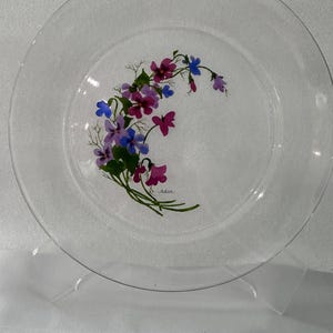 May include: A clear glass plate featuring a hand-painted crescent-shaped arrangement of purple, magenta, and blue flowers with green stems and leaves. The design is positioned off-center, and a small signature is visible near the bottom of the floral cluster.