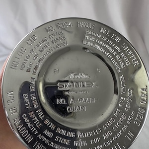 May include: A circular silver-toned metal base of a Stanley vacuum bottle featuring embossed text including 'Aladdin Industries, Inc.', 'Nashville, TN', 'No. A-944DH', 'Quart', and instructions for preheating with hot water or cooling with cold water.