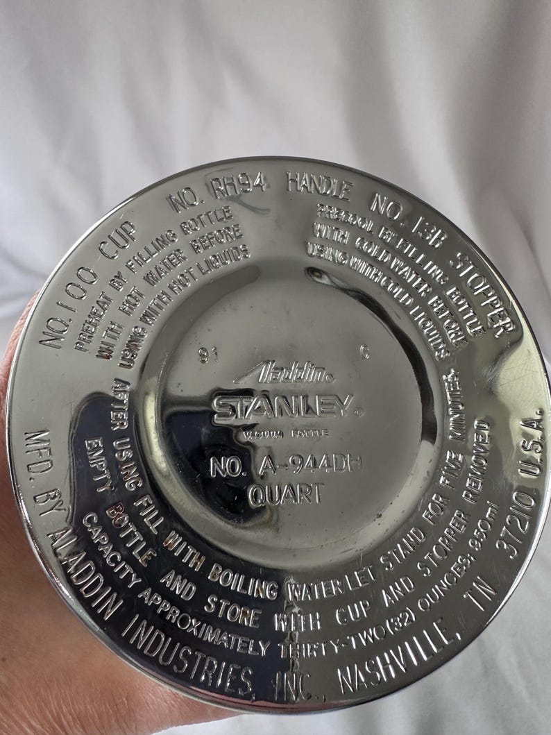 May include: A circular silver-toned metal base of a Stanley vacuum bottle featuring embossed text including 'Aladdin Industries, Inc.', 'Nashville, TN', 'No. A-944DH', 'Quart', and instructions for preheating with hot water or cooling with cold water.