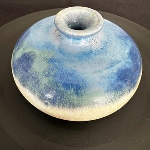 May include: A low-profile ceramic vase with a wide, rounded body and a small, centered circular opening. The surface features a blended glaze of deep blue, light blue, and seafoam green, transitioning to a smooth, creamy off-white base.