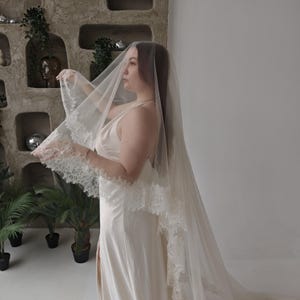 May include: A sheer, ivory-colored wedding veil with delicate lace trim. The veil is long and flowing, creating a soft, romantic look. The bride is wearing a matching ivory slip dress. The background features decorative wall niches and potted plants.