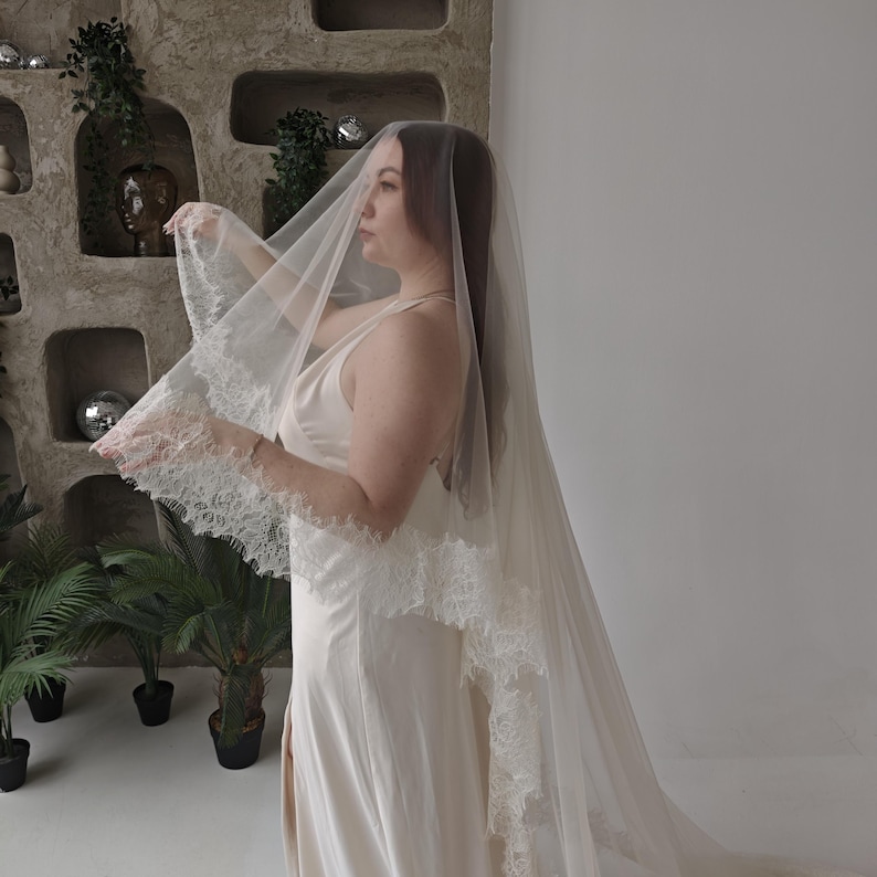 May include: A sheer, ivory-colored wedding veil with delicate lace trim. The veil is long and flowing, creating a soft, romantic look. The bride is wearing a matching ivory slip dress. The background features decorative wall niches and potted plants.