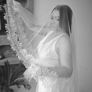 May include: A bride in a white wedding dress and veil. The veil is sheer and has a delicate lace trim. The dress is sleeveless with a V-neckline. The image is in black and white.