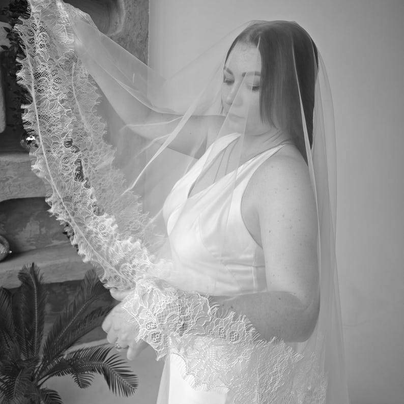 May include: A bride in a white wedding dress and veil. The veil is sheer and has a delicate lace trim. The dress is sleeveless with a V-neckline. The image is in black and white.