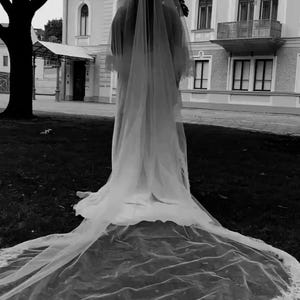 May include: A grayscale image of a person wearing a long, flowing wedding veil. The veil cascades down, forming a train with a delicate lace trim. The background features a building and a tree.