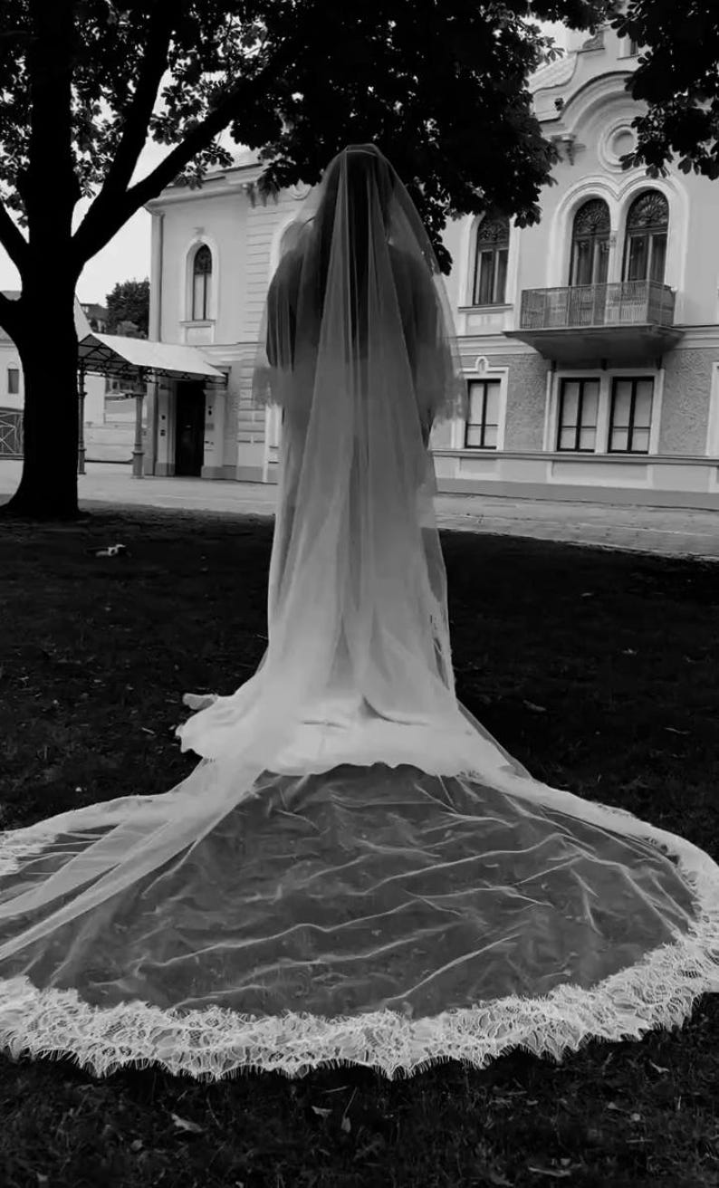 May include: A grayscale image of a person wearing a long, flowing wedding veil. The veil cascades down, forming a train with a delicate lace trim. The background features a building and a tree.
