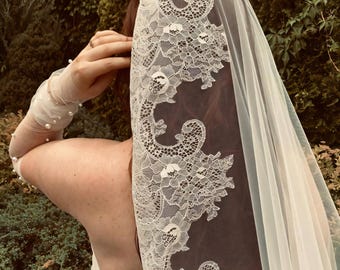 May include: A white wedding veil with intricate lace detailing. The veil is draped over the head and shoulders, with the lace forming a decorative border. The background features lush green foliage, creating a soft, romantic setting.