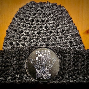 May include: A black and white crocheted beanie with a black circular patch featuring a white moth design. The patch has a white skull and wings with floral details.