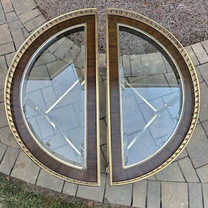 May include: Two half-circle glass-topped tables. Each table has a dark wood frame with a gold trim and a gold metal base. The tables are placed side by side, forming a full circle. The tables are on a stone surface.