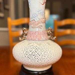 May include: A white ceramic vase with a pink and gold design. The vase has a dragon design on the top and a cut-out pattern on the bottom. The vase is sitting on a black wooden base.